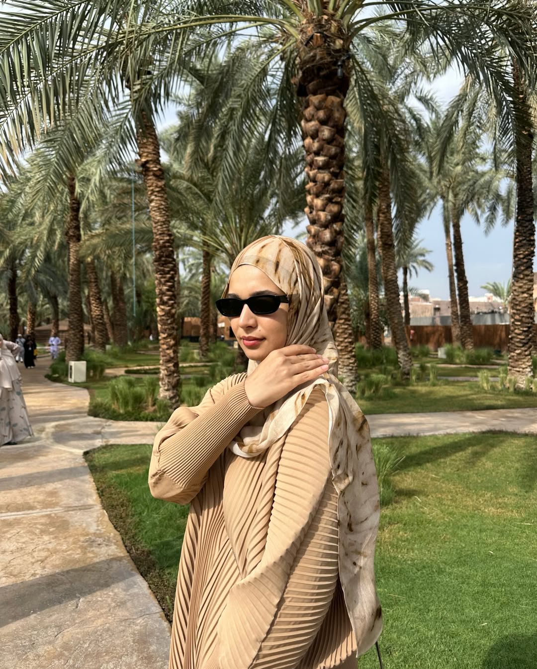 Woman in beige outfit with sunglasses and hijab standing in a palm tree-lined park.